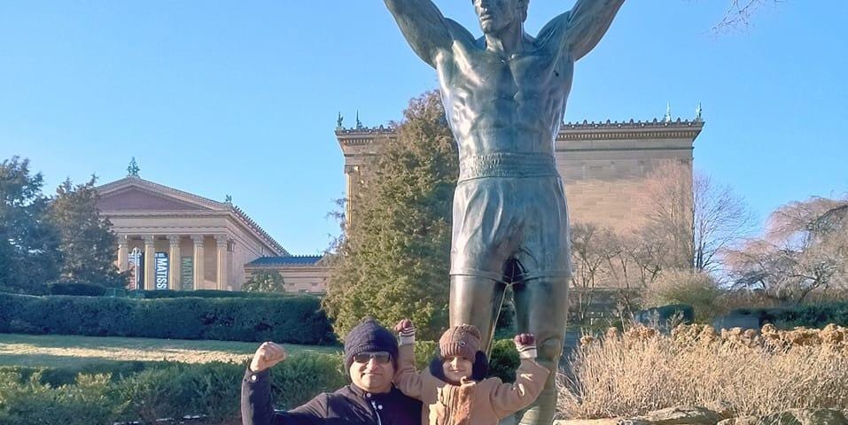 Man carrying a child in front of Rocky Balboa Statue in Philadelphia.