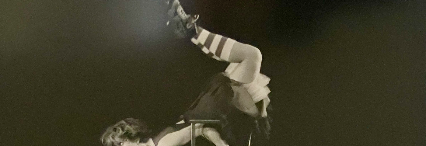 A black and white photo of a short-haired dancer rolling across a stage upside-down on an office chair. She is wearing calf-high, horizontally striped socks.