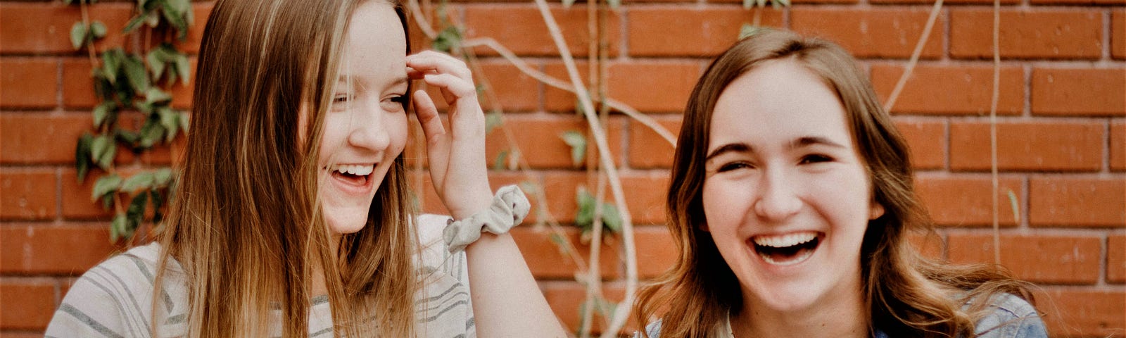 Two teenage girls smiling and laughing in front of red brick wall