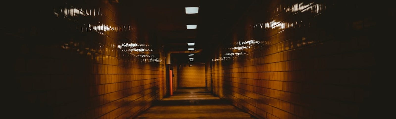 A dark narrow corridor showing a strip of dim lights overhead and tiled walls