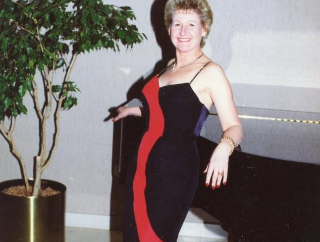 Image of the author in a black dress with a red stripe down the front. She is smiling, and leaning back against a black piano. The photo was taken at a business event, about 28 years ago when she was 52 years old.