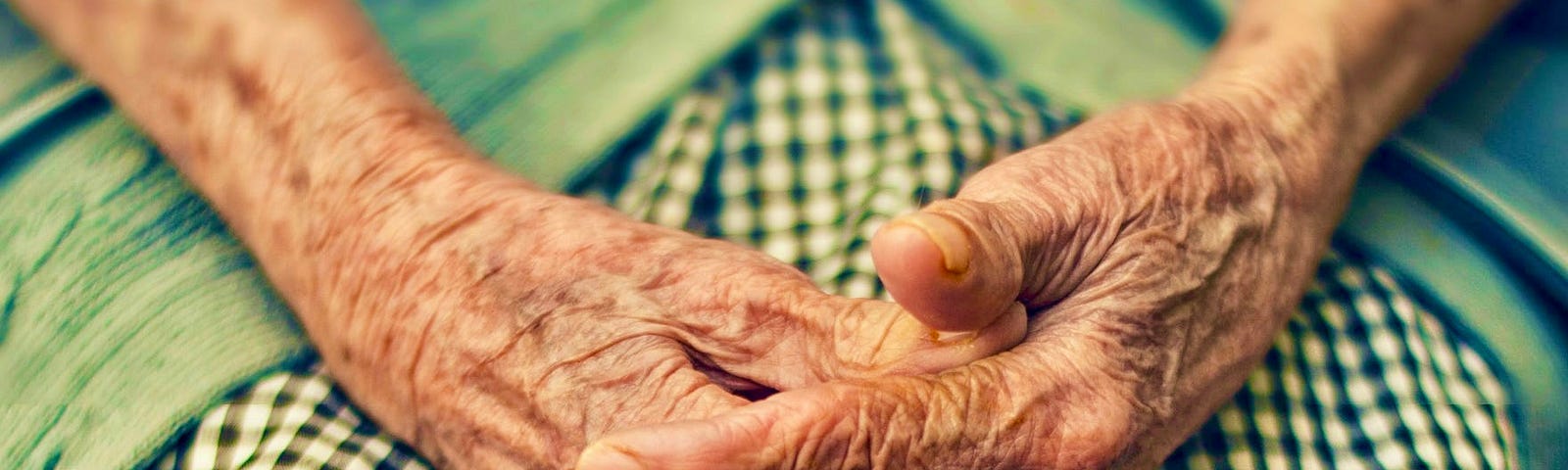 An elderly lady’s hands are clasped in her lap, atop a gingham dress.