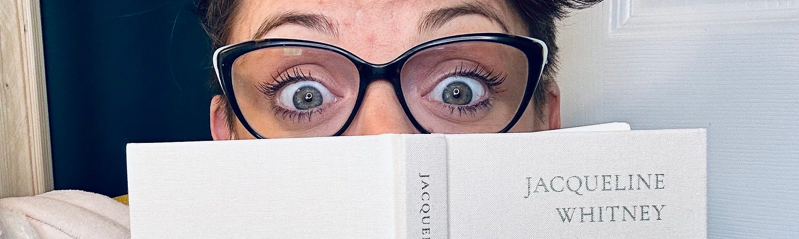 Photo of GiGi hiding her face behind a book titled, “ALL THAT YOU DESERVE”, with excited eyes peeking from behind the top.