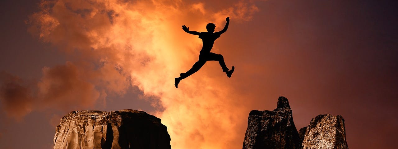 A silhouette of a person leaping from one mountaintop to another. The background is orange sky.