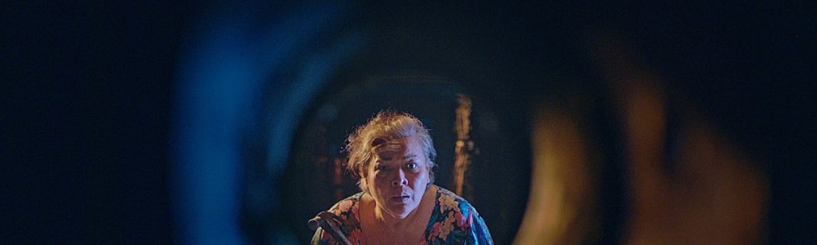 An older woman with mussed hair and wearing a housedress holds a hammer and peers through a pipe.