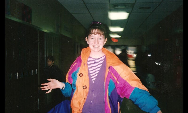 The author as a teenager, with greasy bangs, wearing a purple-and-white-striped shirt under a purple cardigan under a neon multi-colored coat in a middle school hallway.
