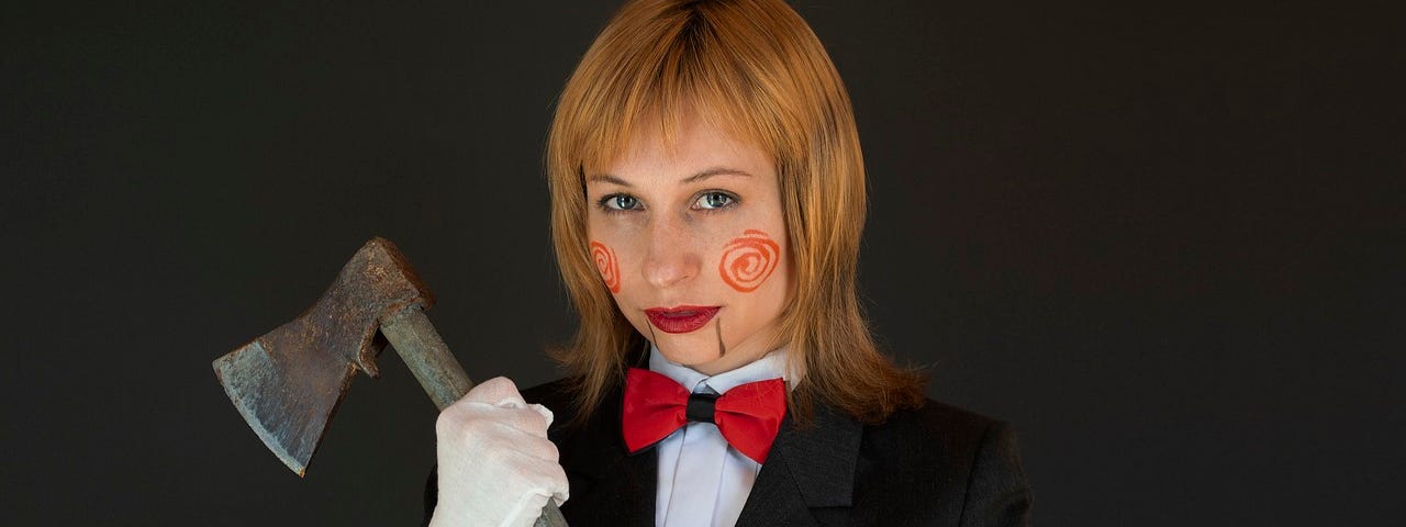 Image has a black background and shows a woman with reddish blond hair, wearing a black blazer, with a white shirt under it and a red bow tie, holding an axe in her hands.