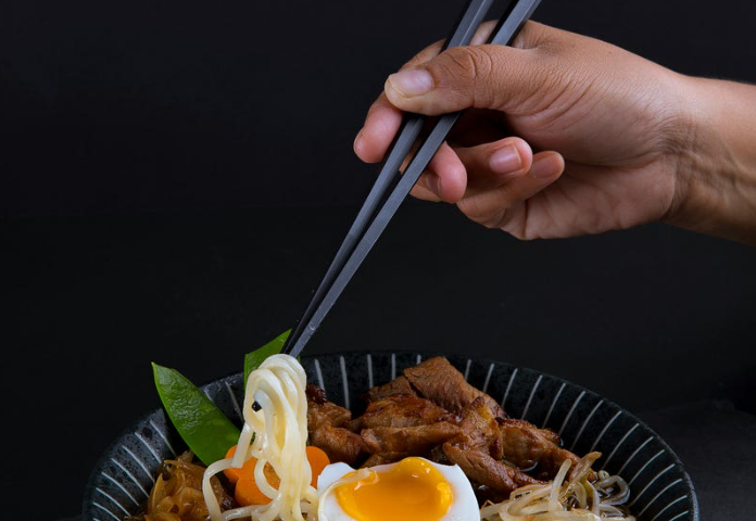 The picture shows a bowl of Japanese noodle with tofu, pork, peas, corns, green vegetable, with someone picking the boiled noodle up with chopsticks.