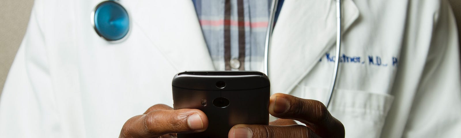 The torso of a Black doctor wearing a flannel shirt, stethoscope, and a white physician’s coat while holding a cell phone with both hands