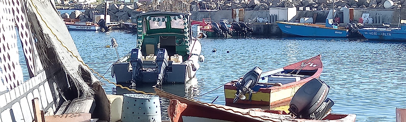 the image is that of a fishing bay in a typical Caribbean island; there are small artisinal boats in the water and on shore and beside them are bigger boats; in the foreground in s fisherman’s shack restaurant and bar.