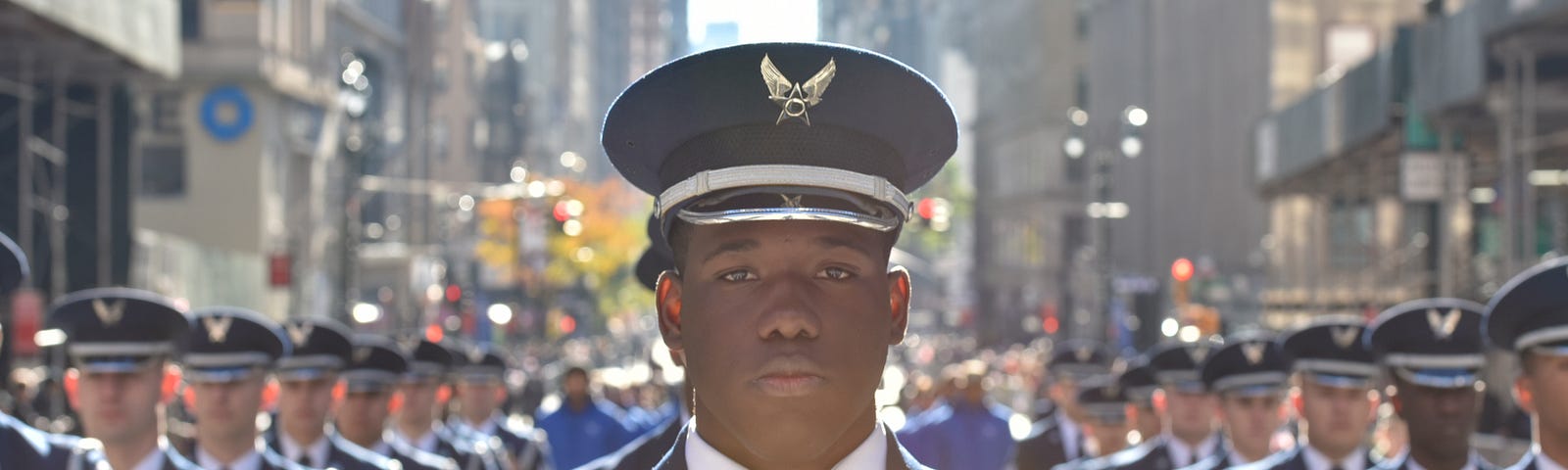 A young man with dark brown skin wears a United States Air Force uniform and stands in the streets of New York City flanked by fellow airmen for the Veterans Day Parade in 2018.
