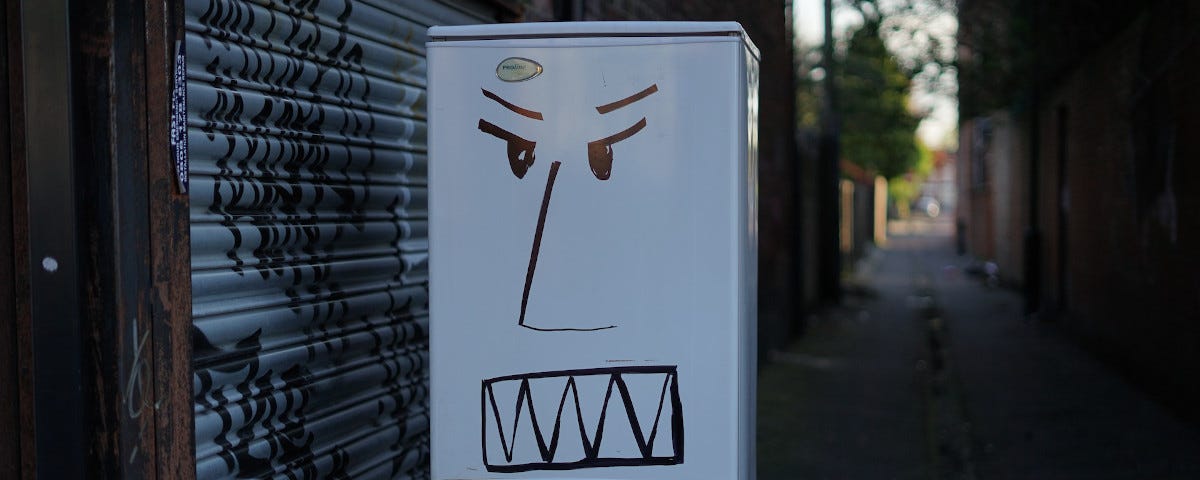 A white fridge-freezer abandoned in an alleyway, outside the door to a lock-up. An angry face has been drawn with black marker on the fridge door, and a hashtag — Fridges have feelings too — has been written on the freezer door.