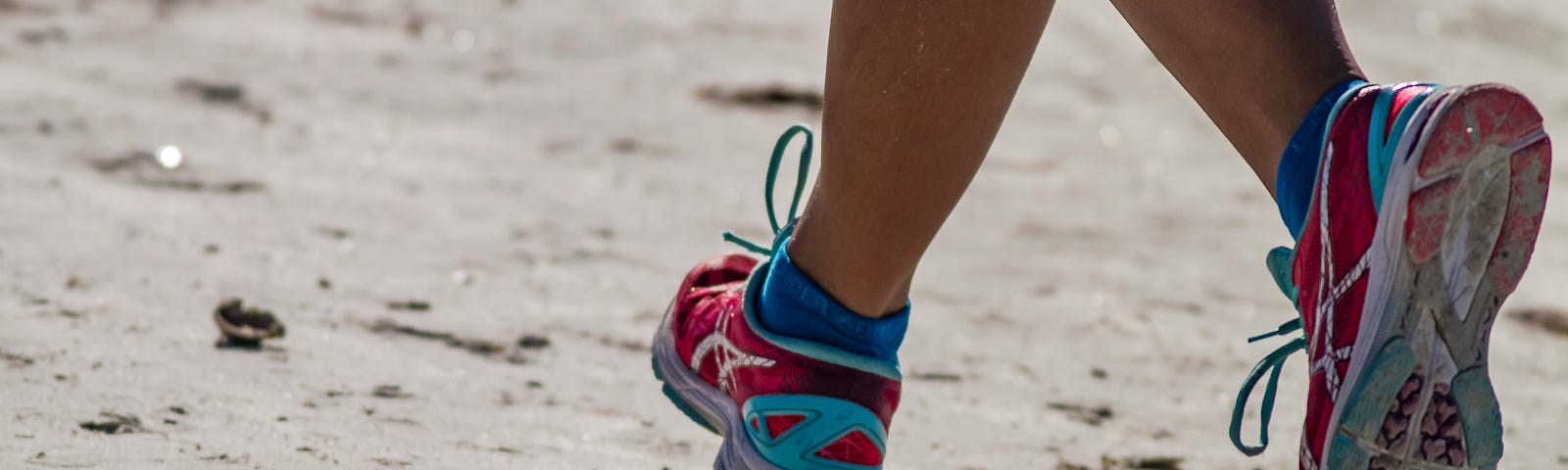 The legs of a woman shown running on a beach wearing pink and blue tennis shoes.