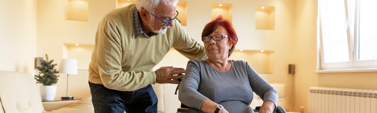 Older Asian woman in wheelchair looking at an older White man leaning over her shoulder to talk to her. Photo by ProfessionalStudioImages/Getty Images