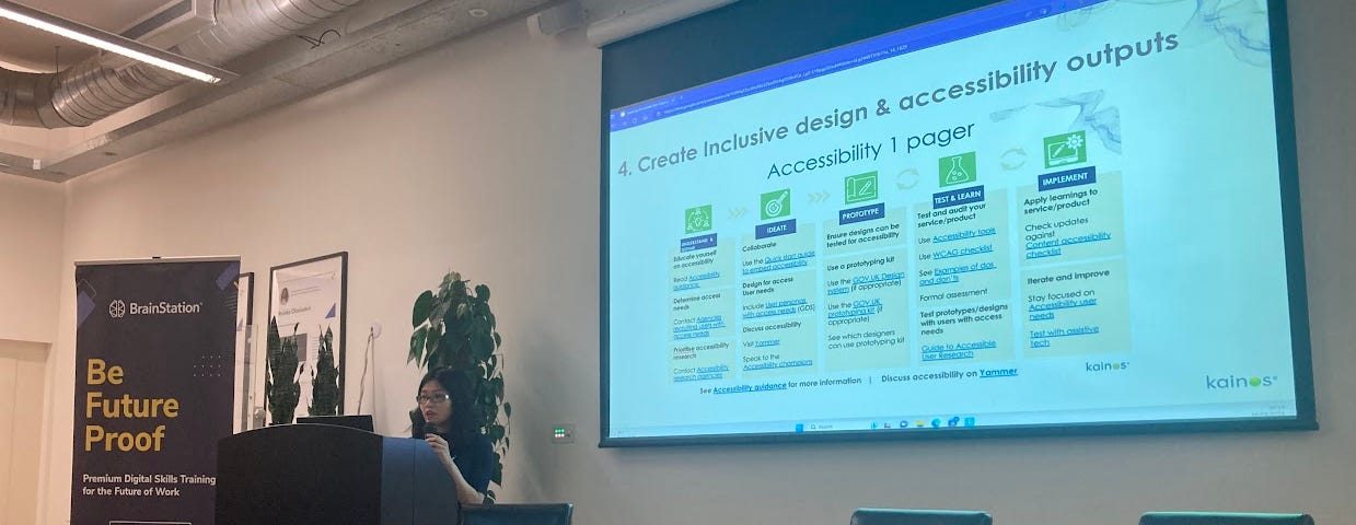 Christina Lai, an east asian woman of short stature presents inclusive design and accessibility outputs on a projector, next to podium and 3 empty stools. Banner beside her reads ‘BrainStation. Be Future Proof’