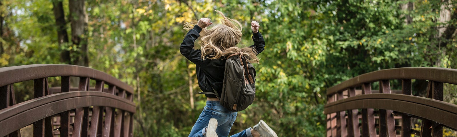 Wooden bridge going into forest with bright green trees and sun peaking through. The bridge has dark brown arched rails with Vs alternating up and down, the path is light brown wooden planks. Towards the middle of the bridge is a young blonde haired girl jumping in celebration into the air. Her legs are both bent at the knees, her arms are pumped into the air, and her hair is blowing in the wind. She is in blue jeans, a dark top, and has a brown backpack.