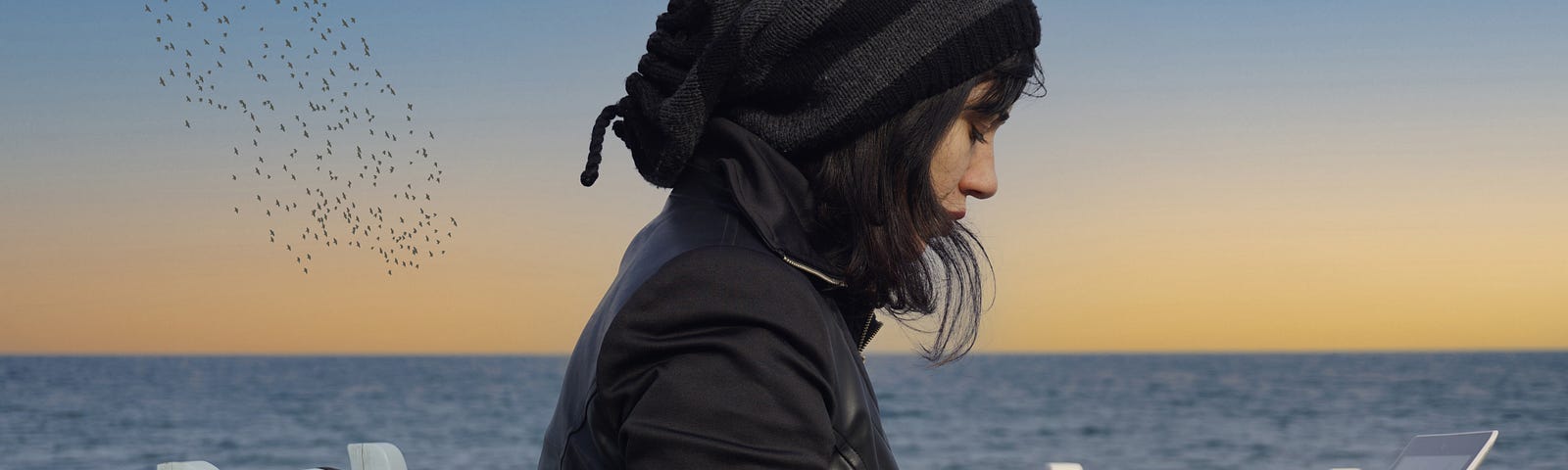 A woman wearing all black types at a table on the beach with a body of water in the background.