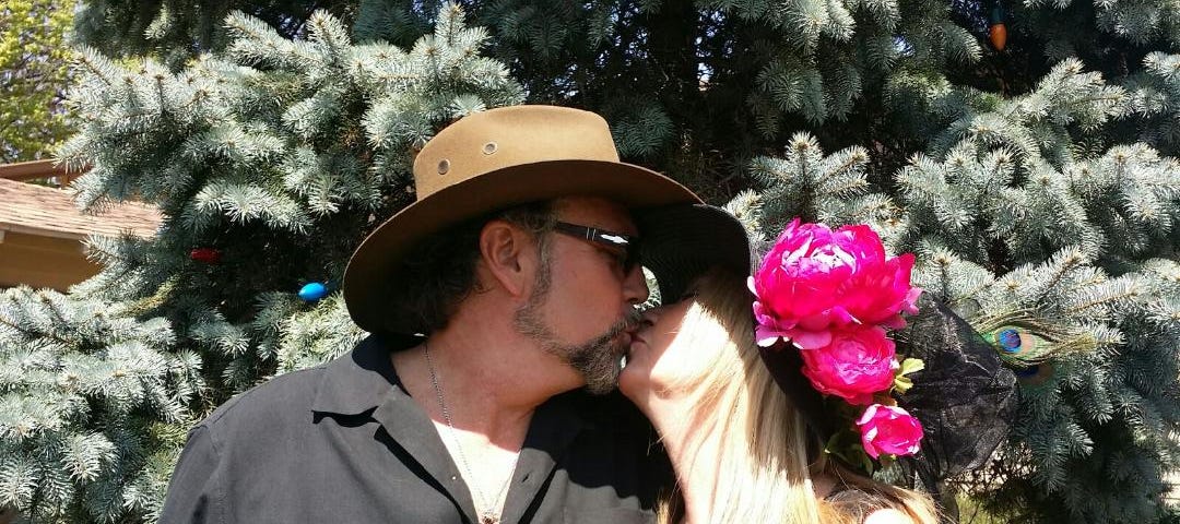 Man and woman kissing at a Kentucky Derby party, he with a brown Stetson and she with a big black hat decorated with fuchsia pink peonies.