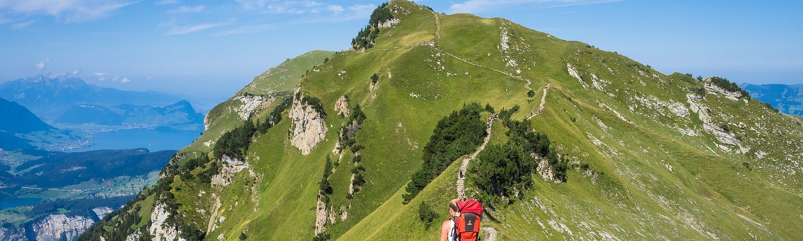 A woman with a red backpack going up a sinuous dirt mountain path, with more people higher above. The sides of the mountain are covered in grass and the clear blue sky is in the background.