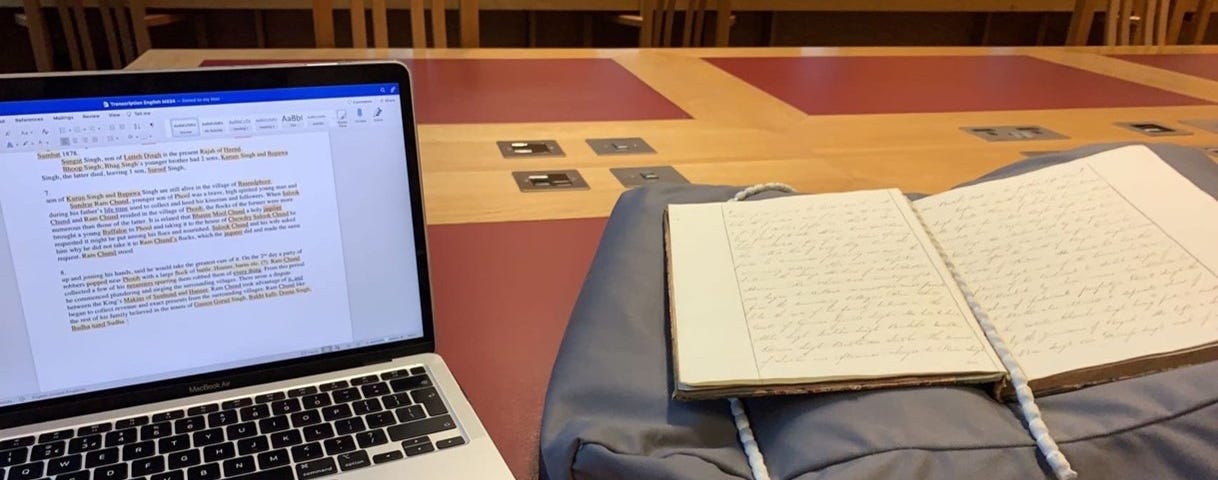 A desk in the Elsevier Reading Room featuring a laptop (left) displaying a word document of the typed transcript — with many highlighted sections — alongside a manuscript (right), supported by a pillow and held down by snake weights.