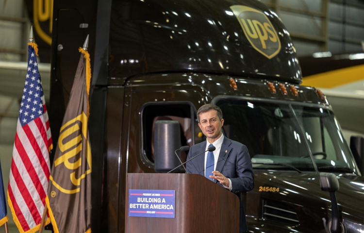 Secretary Buttigieg delivers remarks at a podium with an American flag and UPS truck in the background.