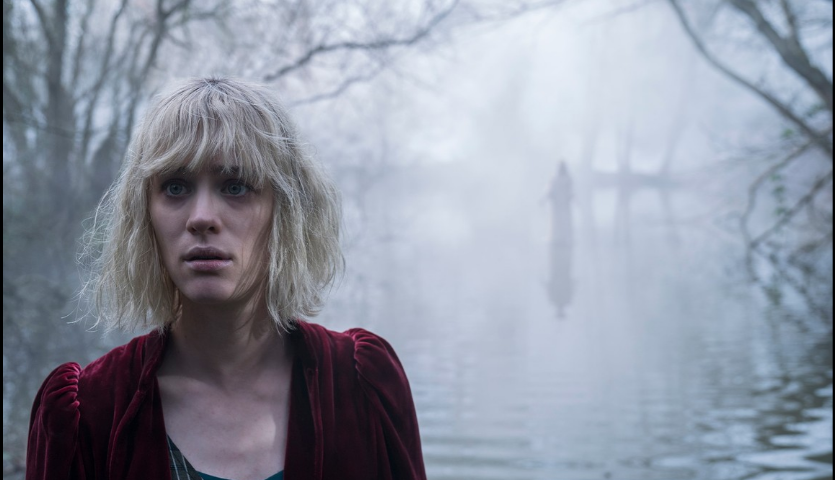 Kate standing in front of the lake, with a ghost standing behind her on the water in The Turning (2020)