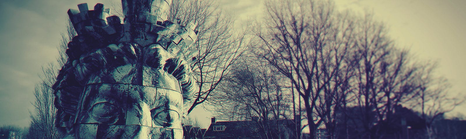 black and white photo of a dilapidated in house behind a set of three bare trees with branches extending upward. In the front left is a large broken statue of a head.