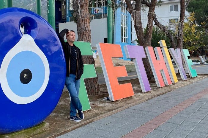Man in winter clothes leaning on large multi colored lettered sign for Fethiye