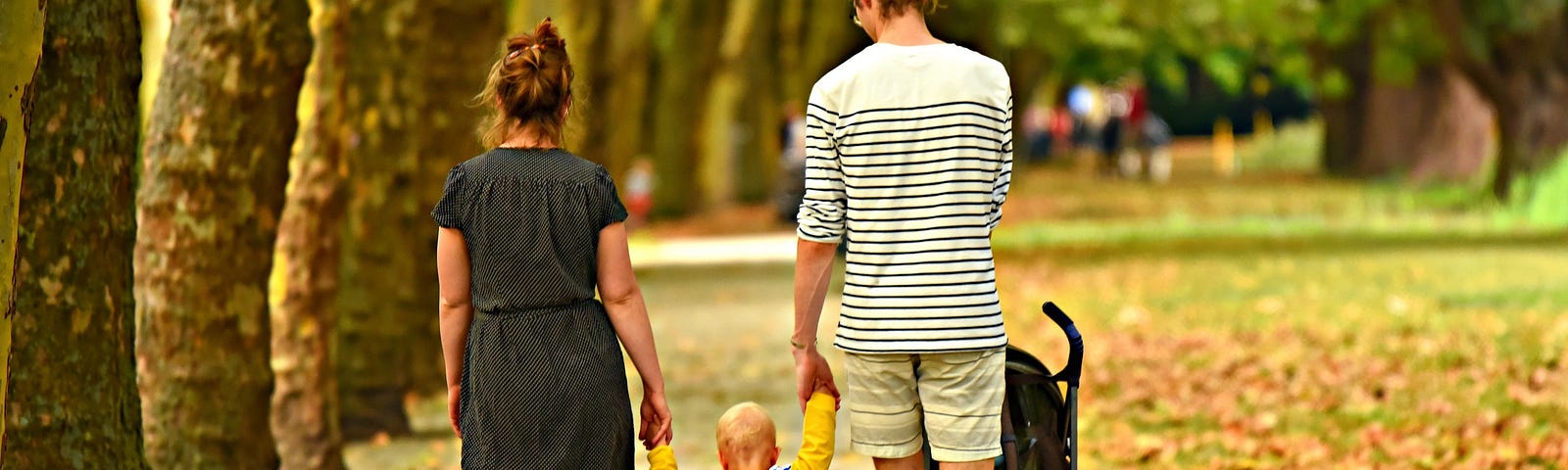 Young woman and man walking in a park in the fall and holding hands with a toddler in a yellow jacket walking between them.