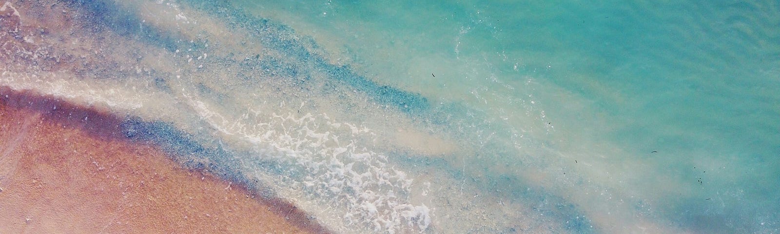 A pale turquoise sea crashes against a beach of peachy pink