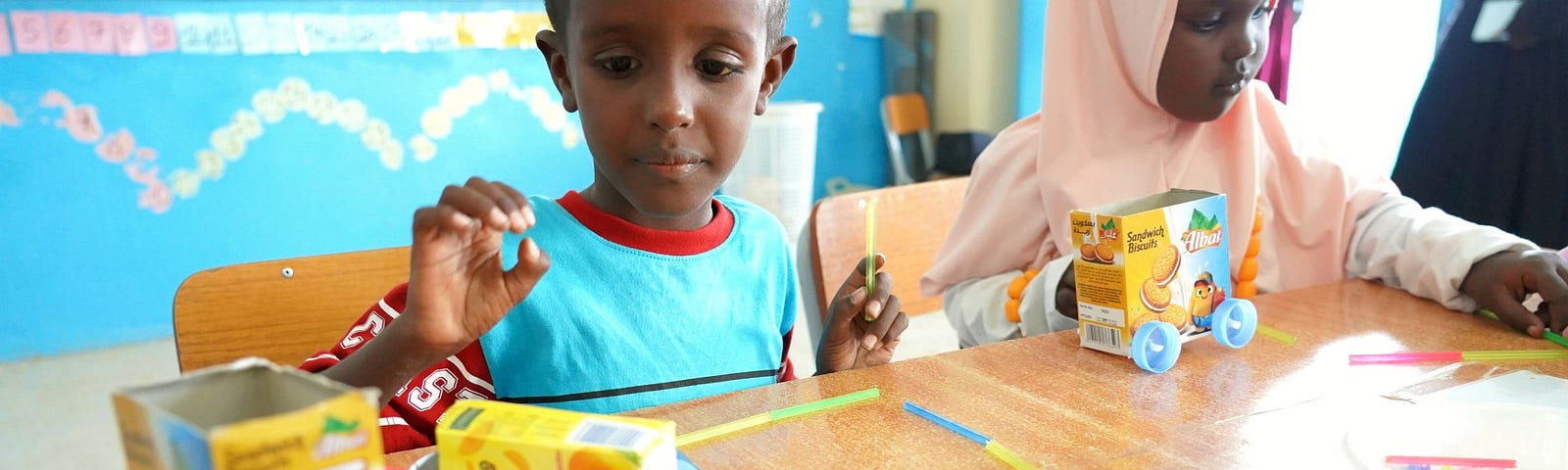 A boy and girl sit at their work stations at school with the toy cars they have built with small recycled cardboard boxes, colorful drinking straws, and little blue wheels.