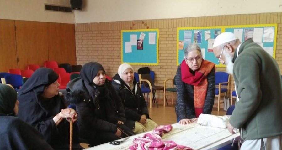 A group of women wearing headscarves are sat at a table listening to an older man and woman talking through the sewing materials in front of them.