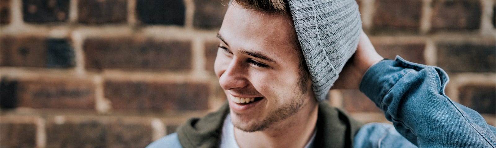 smiling man wearing blue jean jacket and blue knit hat in front of brick wall