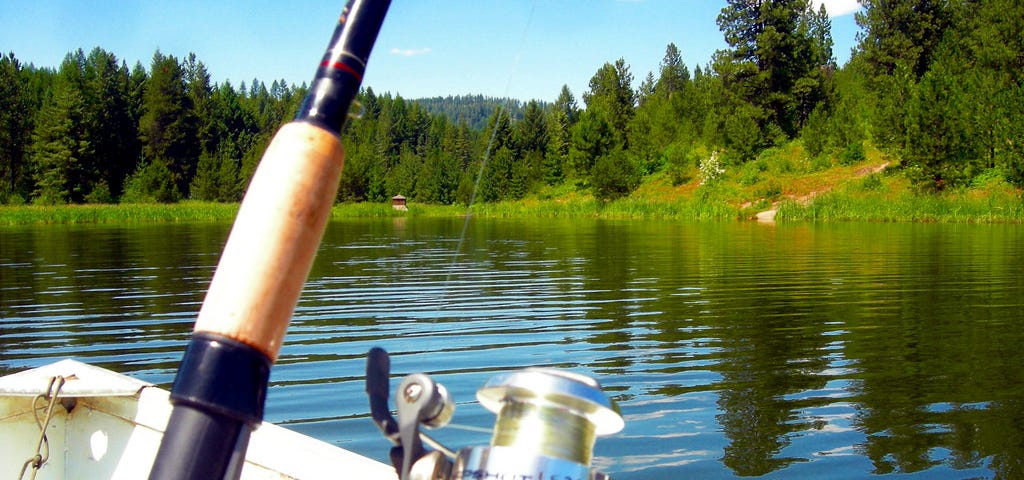 A photograph of a the handle of a fishing pole with a reel in the forground with the bow of the boat just behind in a lake looking at the shorline in the background on a sunny day with blue sky.