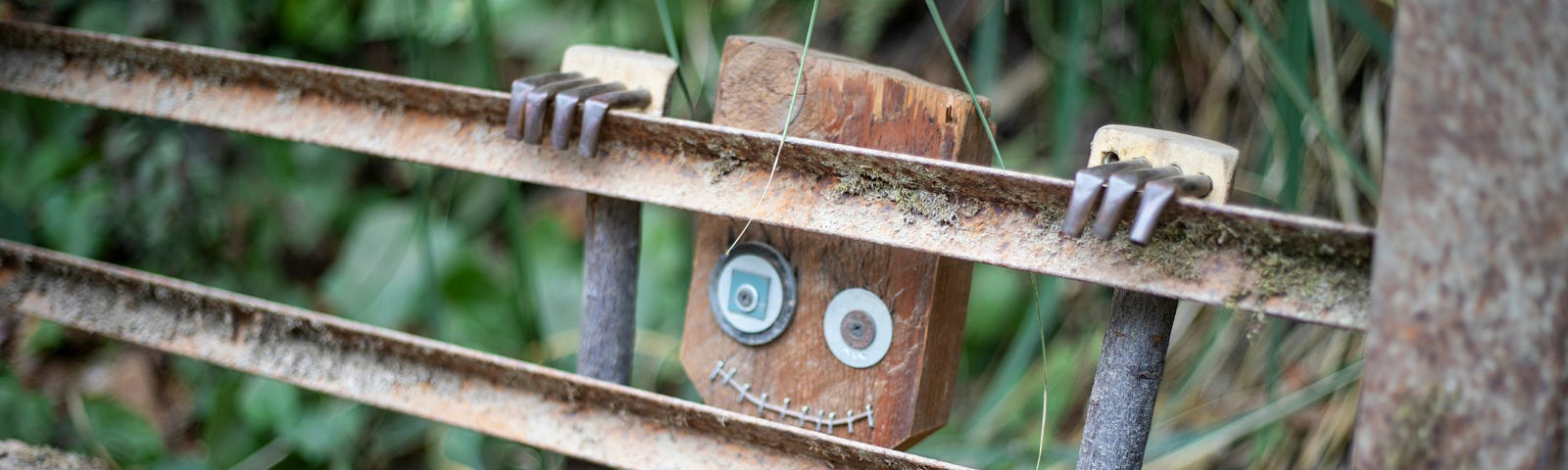 The image shows a small wooden figure resembling a robot with a square head, large round eyes made from washers, and a stitched mouth. The figure is positioned behind rusted metal bars on the side of a bridge or walkway, peering through. The background includes blurred greenery.