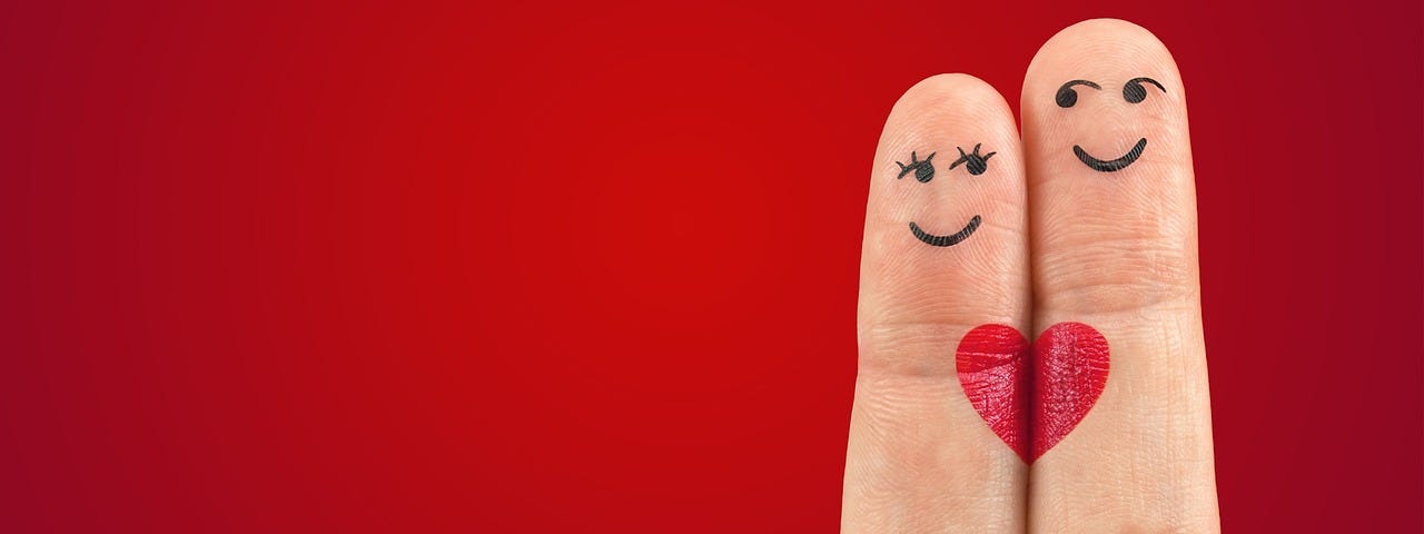 Two fingers close together, the shorter one has female eyes and mouth drawn on it, the taller has male eyes and a smile drawn on it, Each has half a red heart which — side by side — make a whole heart