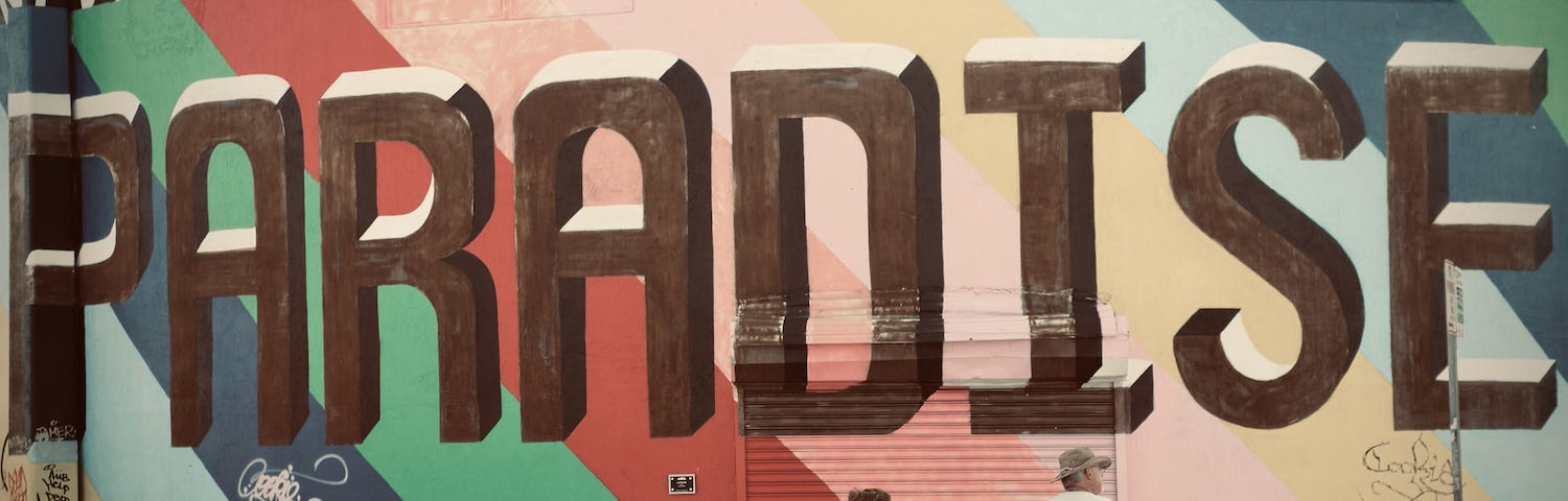 A picture of a couple holding hands in front of a graffiti wall with the word “paradise” in huge brown letters on a background of bright colors.