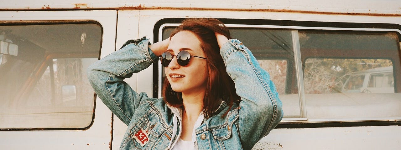 Young girl in sunglasses and a jean jacket, standing in front of vw van, with her hands on her head, smiling at awe at the scene in front of her. (A sunset perhaps?)