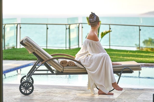 Woman sits on deck lounger, wears white peasant style dress off shoulders, hair up, bare feet. She’s facing away at a blue sea view. holiday