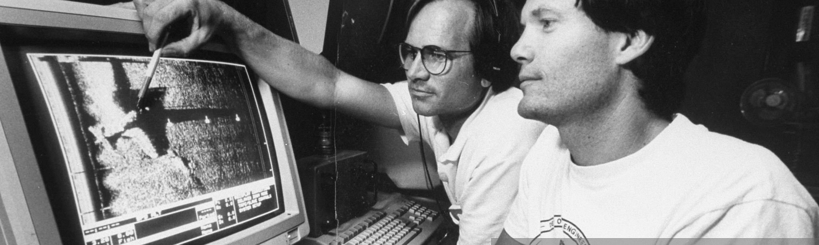 A black and white photo of two explorers of Deep See Co., a company that searches for sunken ships and planes in the Bermuda Triangle. The photo shows the explorers in a boat, looking at a computer screen that displays a sonar image of one of the five Navy Avenger Torpedo Bombers that they found. The bombers have been lost in the Bermuda Triangle since December 1945. The photo was taken by Acey Harper and Getty Images.