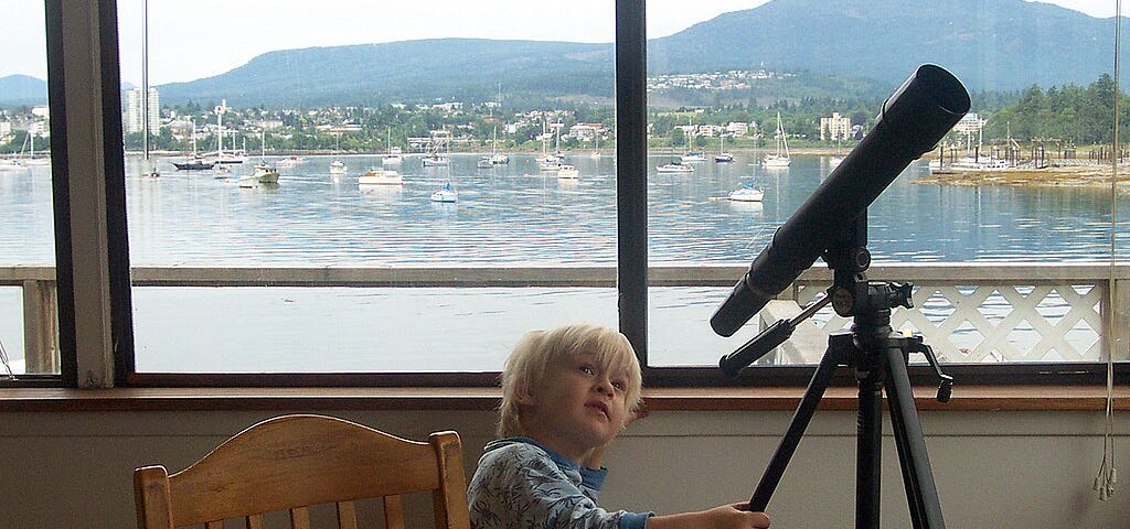 A child in pajamas looks at a telescope on a tripod. Behind him, small boats can be seen on the lake.