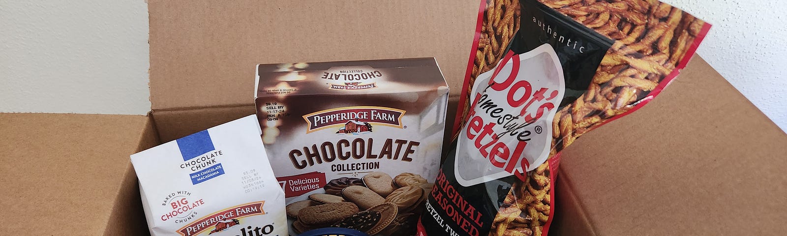 An open box reveals two different kinds of Pepperidge Farm cookies, a bag of Dot’s Homestyle Pretzels, and a small can of Planters Deluxe Mixed Nuts.