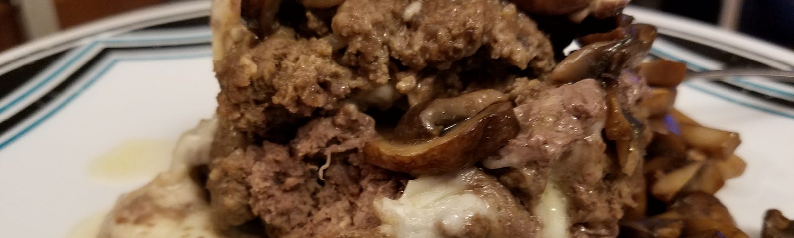 A ball shaped hamburger patty that had been stuffed with cheese and cooked in a presure cooker, then topped with sauteed mushrooms on a white plate.