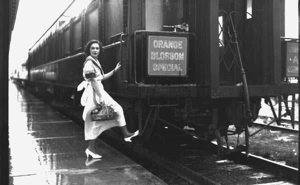 A 1930s black and white photo of a woman with her foot on the step on the last car of a train. Her hand is holding the handrail. She is wearing a white dress and white shoes and carrying a purse.