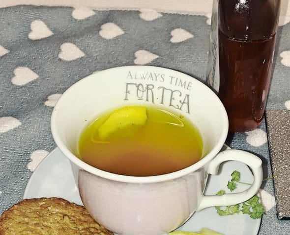 A cup of herbal tea with a slice of lemon and a biscuit on the saucer. The cup has the words ‘Always Time For Tea’ inscribed at the top.