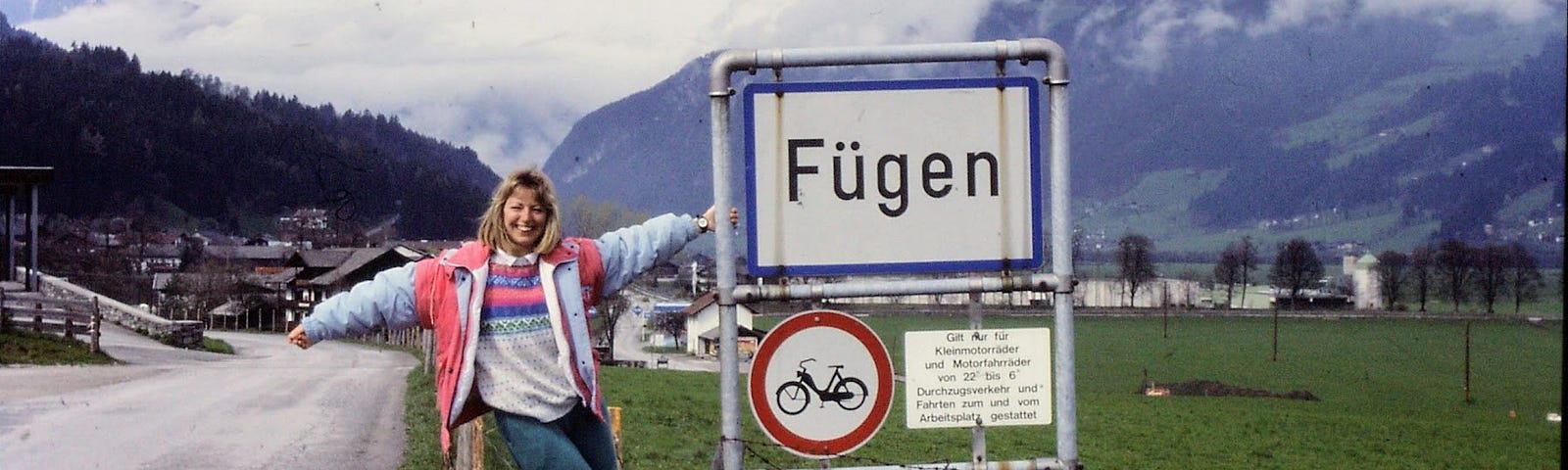 A woman standing on a wooden fence and holding onto a village sign, with a field and mountains in the background.