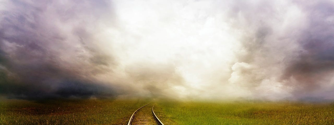 A young child sits looking away into the distance on railway tracks with a teddy bear at their side. The tracks are on grass and disappear into the clouds.