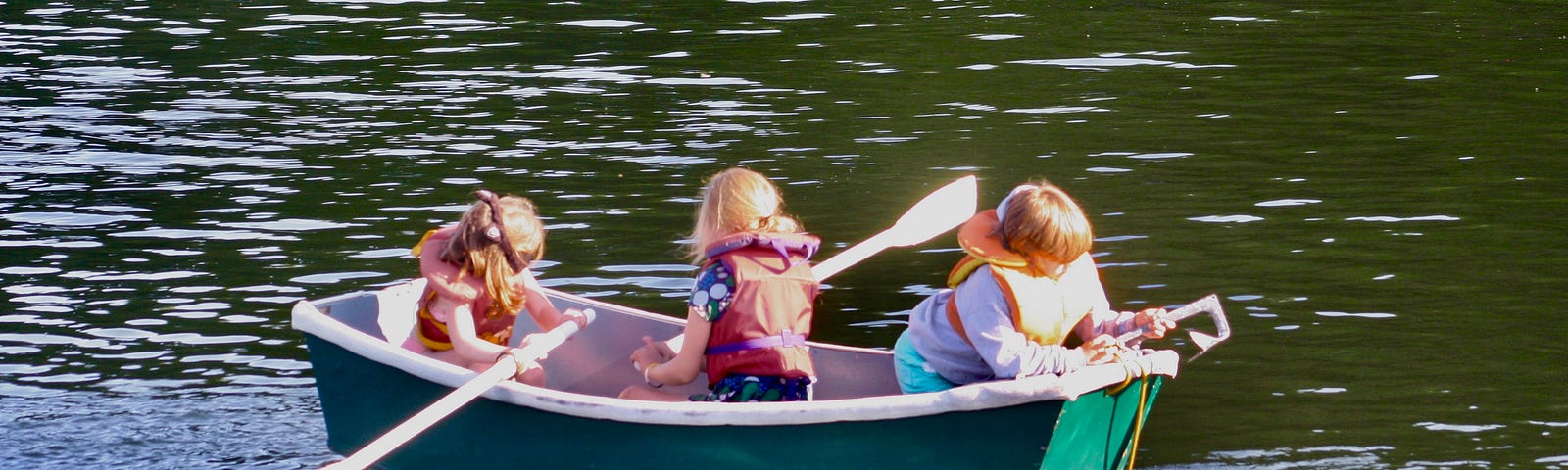 Three children sit in a green rowboat. Two of them are holding oars. One is hanging over the stern of the boat.