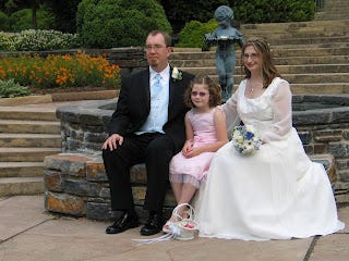 Photo of a man in a tuxedo, a child in a pink dress, and a woman in a bridal gown seated on stone wall.