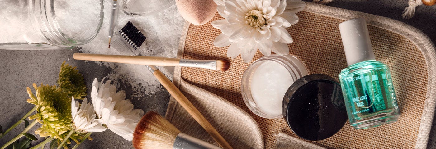 Big and small makeup brushes, spilled bath salts, green and white chrysanthemums, a bottle of transparent blue nail polish, and other beauty products lay flat on a gray table-top and an open beige makeup case.